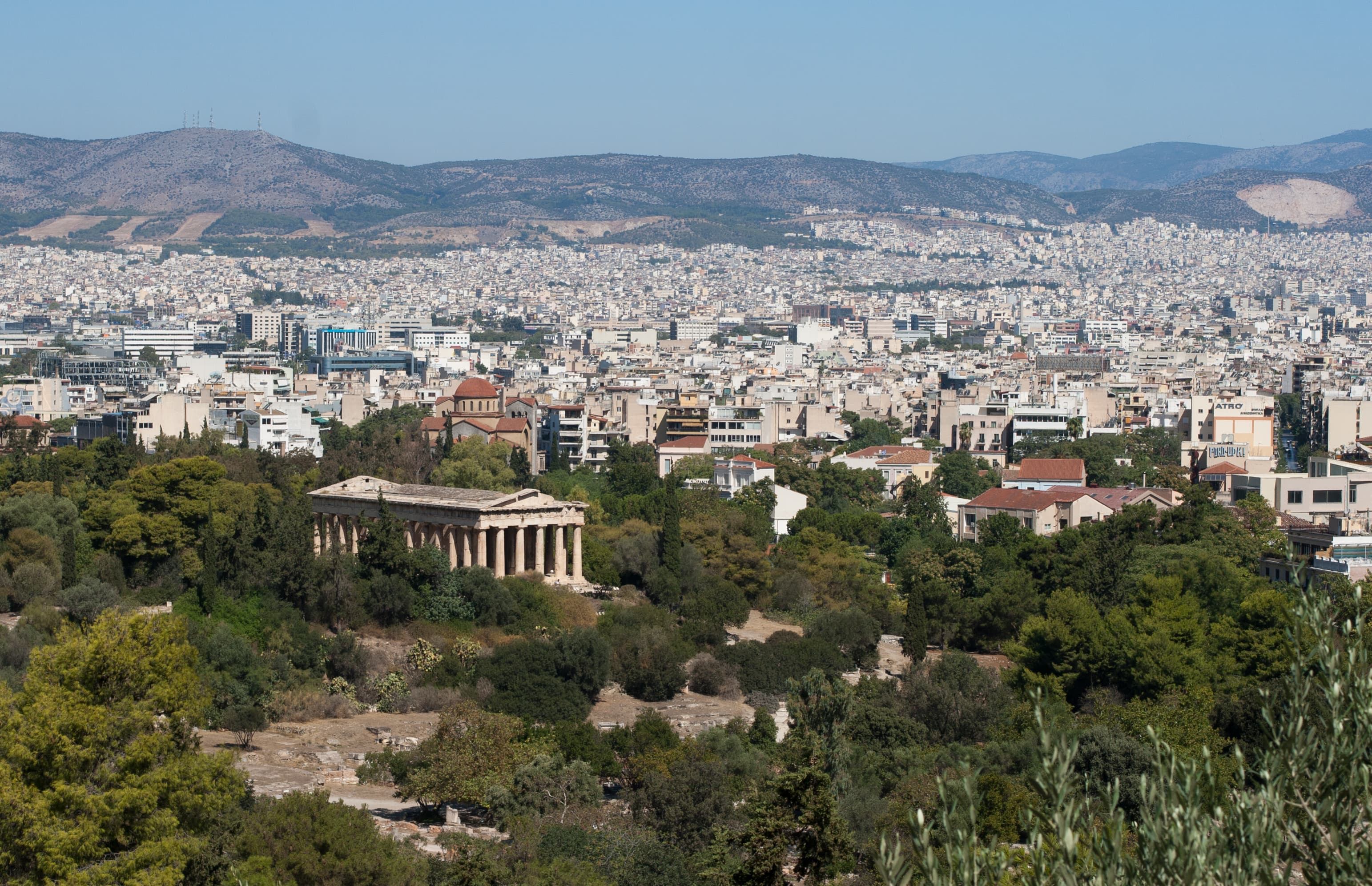 View over Athens from the Acropolis hill, with the Temple of Hephaestus in the Ancient Agora visible among the trees, the city spreading north toward the mountains