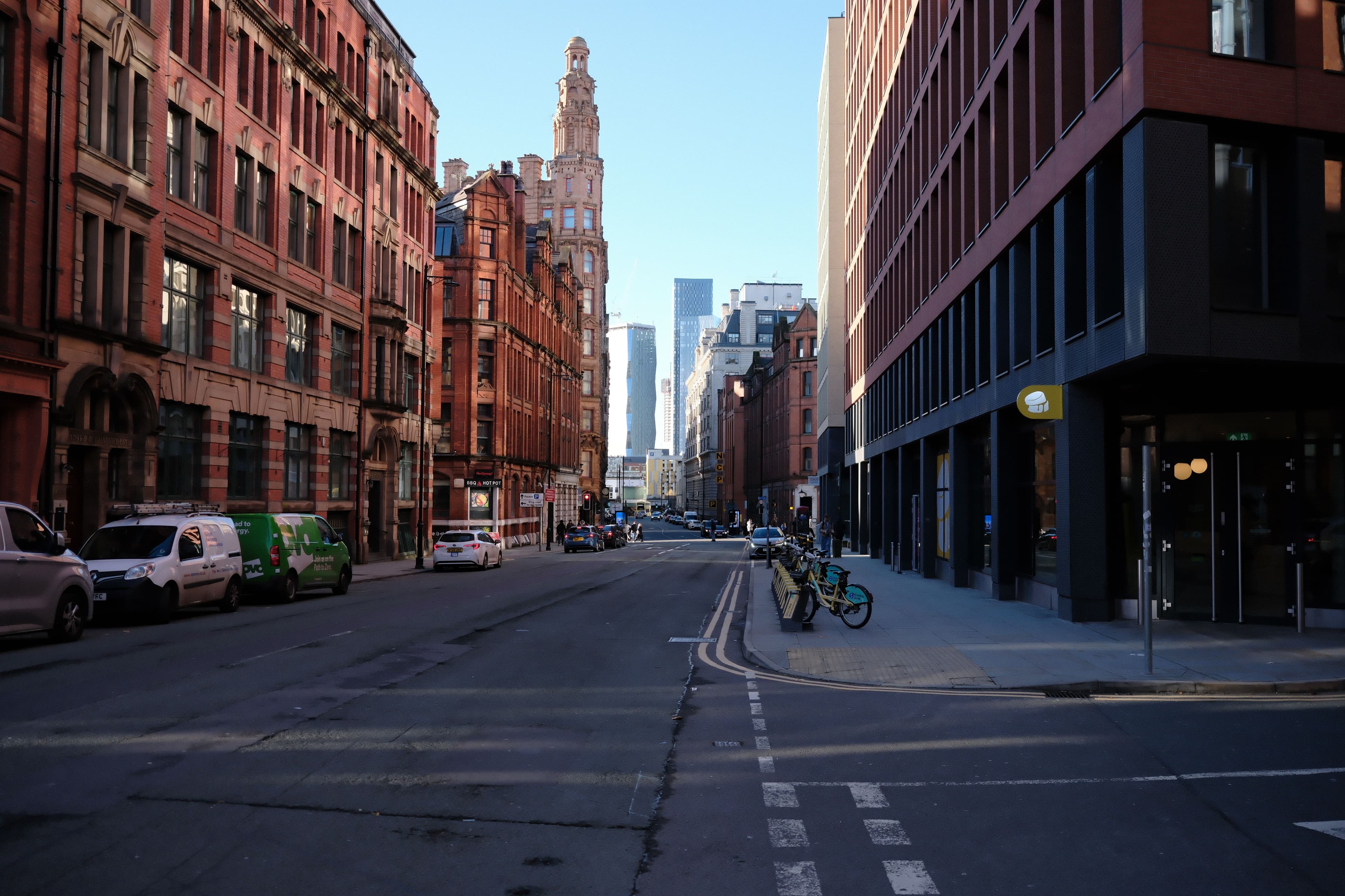 A quiet Manchester city street lined with Victorian red-brick buildings, with the Beetham Tower and modern city-centre towers visible in the distance